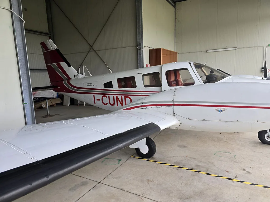 A white Piper Seneca II twin-engine aircraft with red and black stripes is parked inside a hangar, showing its registration I-CUND.