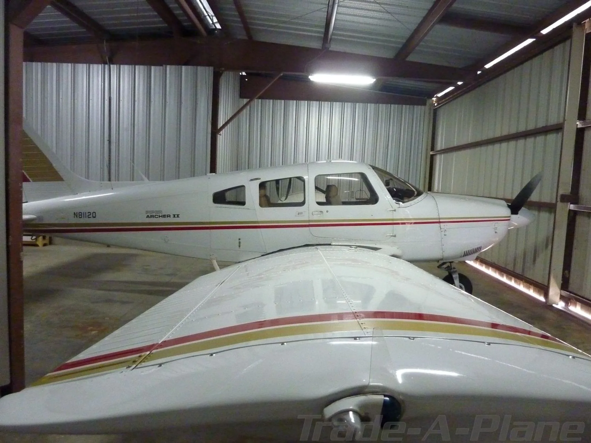 A view from over the wing of a white Piper Archer II aircraft with red and gold stripes, registration N8112Q, in a hangar.