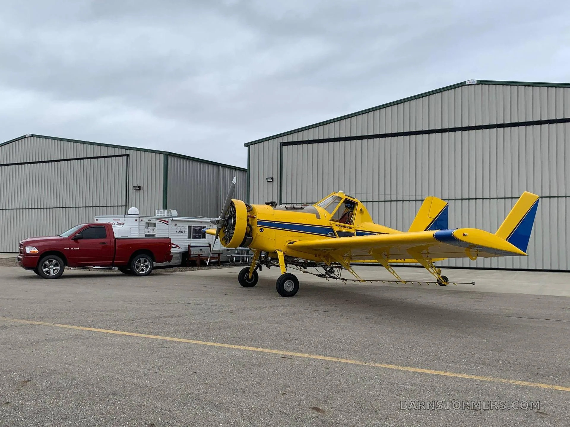 A yellow agricultural aircraft with blue stripes parked on an tarmac next to a red pickup truck and hangars under a cloudy sky.