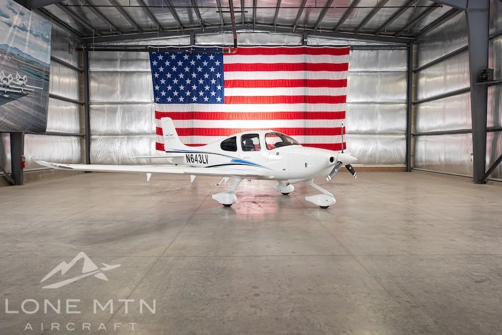 A white Cirrus SR22 aircraft with blue and black stripes, tail number N643LV, parked in a hangar with an American flag in the background.