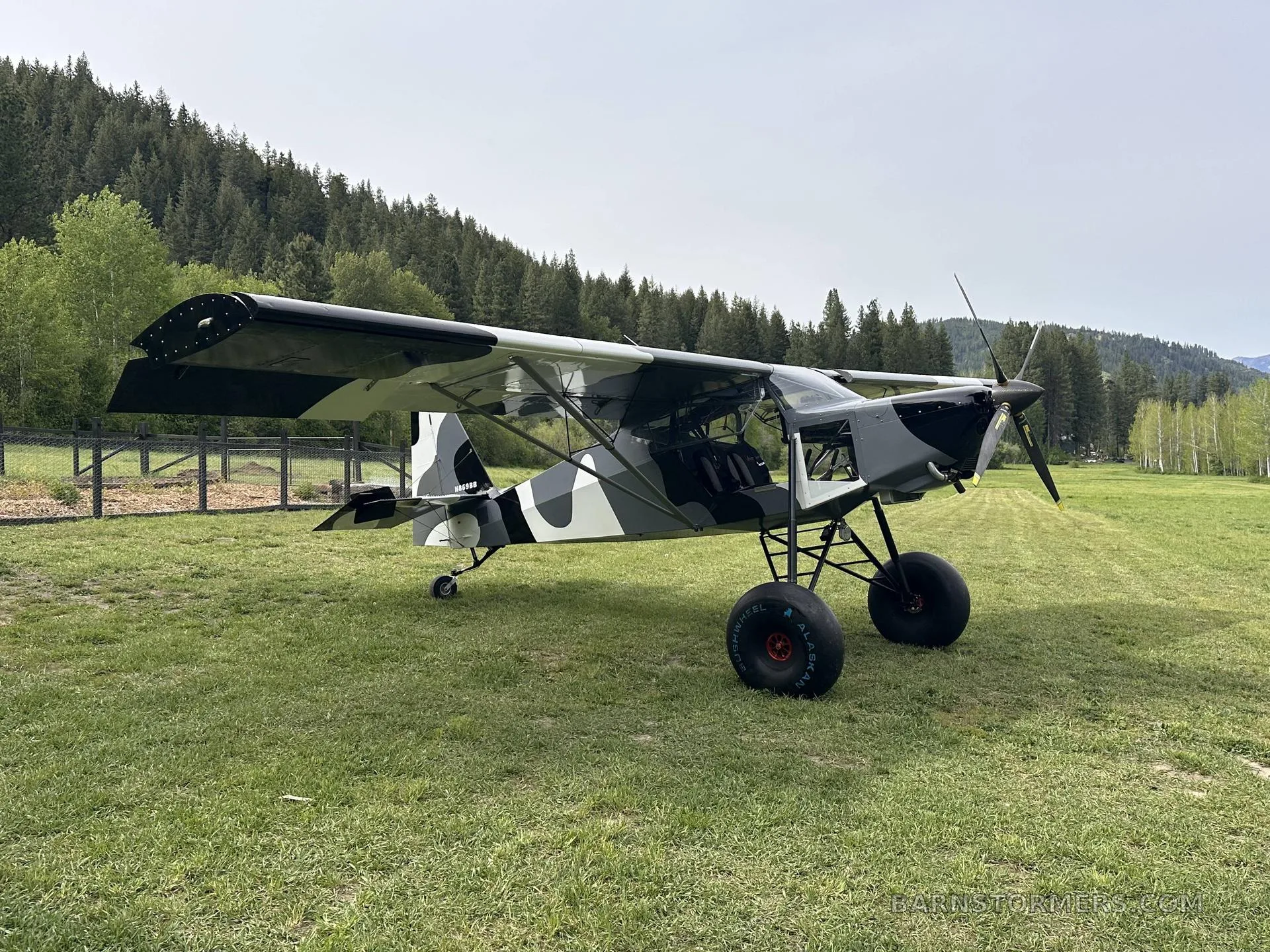 A black, white, and gray camouflage STOL aircraft with large tundra tires is parked on a grassy field, viewed from the front-left.