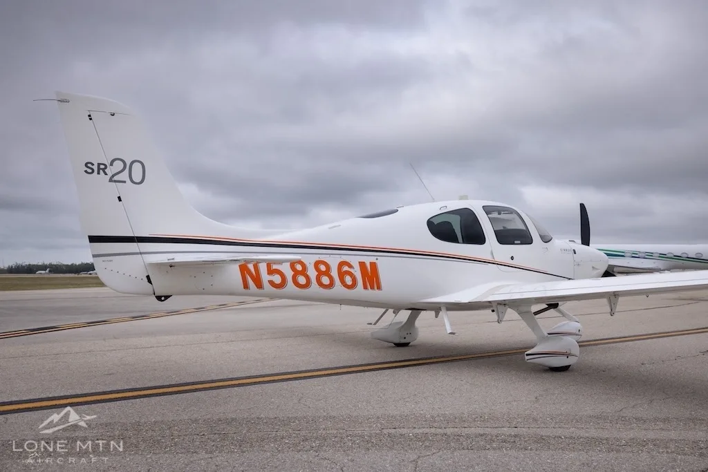 A rear-side view of a white Cirrus SR20 aircraft with orange and black stripes, registration N5886M, on an airfield.