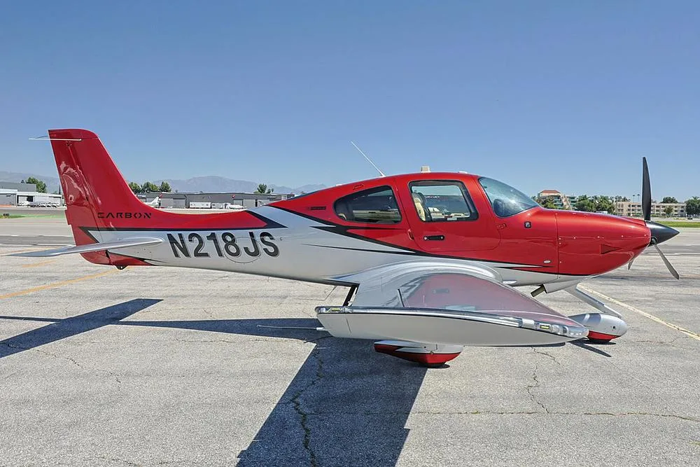 A close-up side view of a red and silver Cirrus SR22 aircraft with registration N218JS parked on a sunny tarmac.