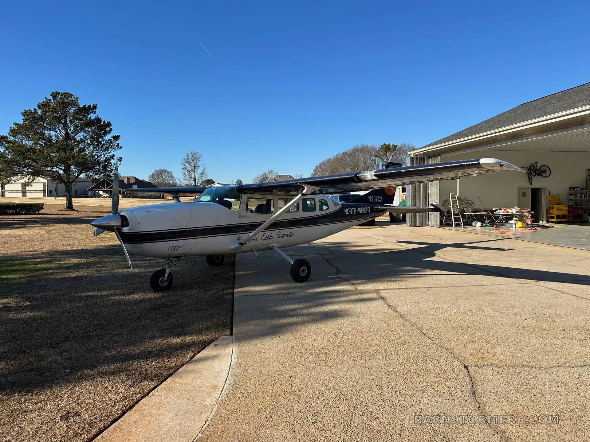 A white and dark blue Cessna 207 aircraft, N207CZ, with "NORTH-WRIGHT" text, parked on a concrete driveway with a house and trees in the background.
