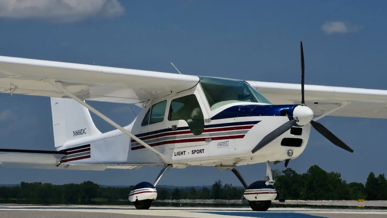 A white Paradise P1 Light Sport aircraft with blue and red stripes on the nose and fuselage, parked on a tarmac under a blue sky.