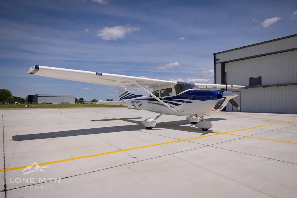 A white and blue Cessna 182 Skylane (N27GH) parked on an airport tarmac with a hangar in the background.