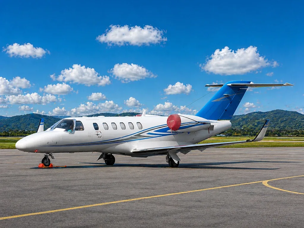 A white business jet with blue and grey stripes and a blue tail is parked on an airport tarmac with mountains in the background.