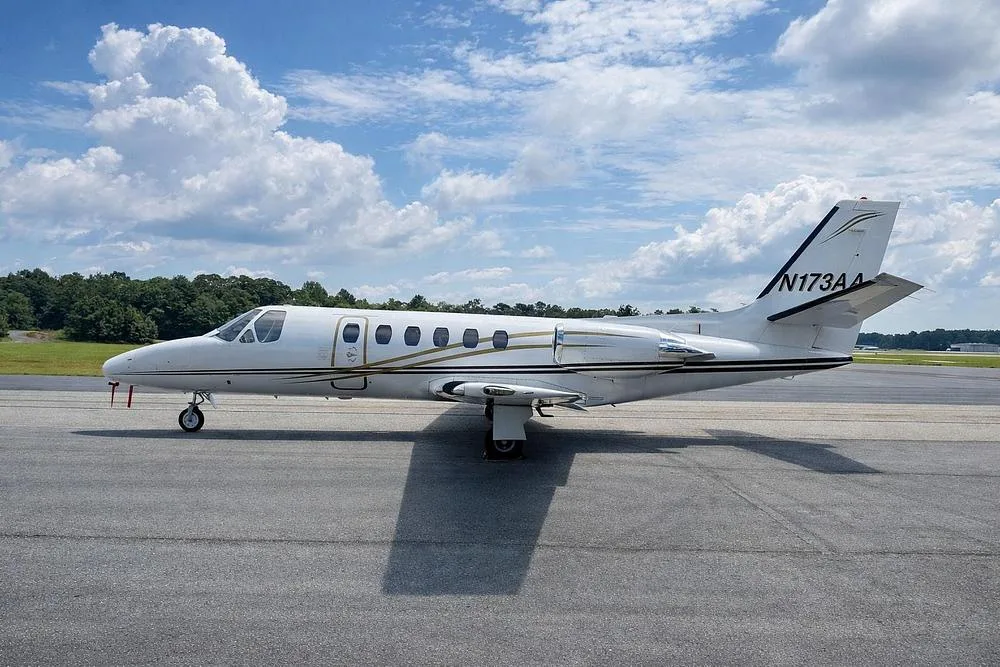 A white business jet with gold and black stripes, tail number N173AA, parked on an asphalt runway under a partly cloudy sky.
