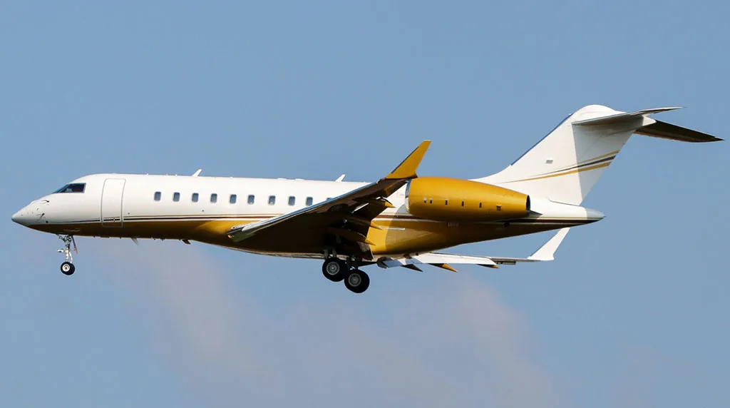 A white and gold Bombardier Global Express business jet with landing gear extended, flying against a clear blue sky.