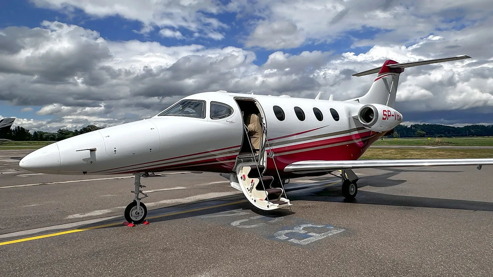A white and red business jet, tail number SP-VHA, is parked on an asphalt tarmac with its door open under a cloudy sky.