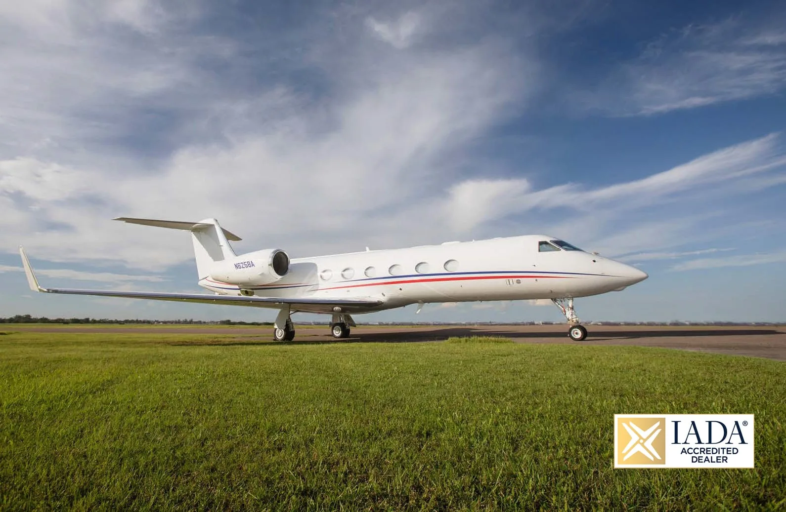 A white Gulfstream G650 business jet with red and blue stripes on the fuselage, parked on a runway with grass.