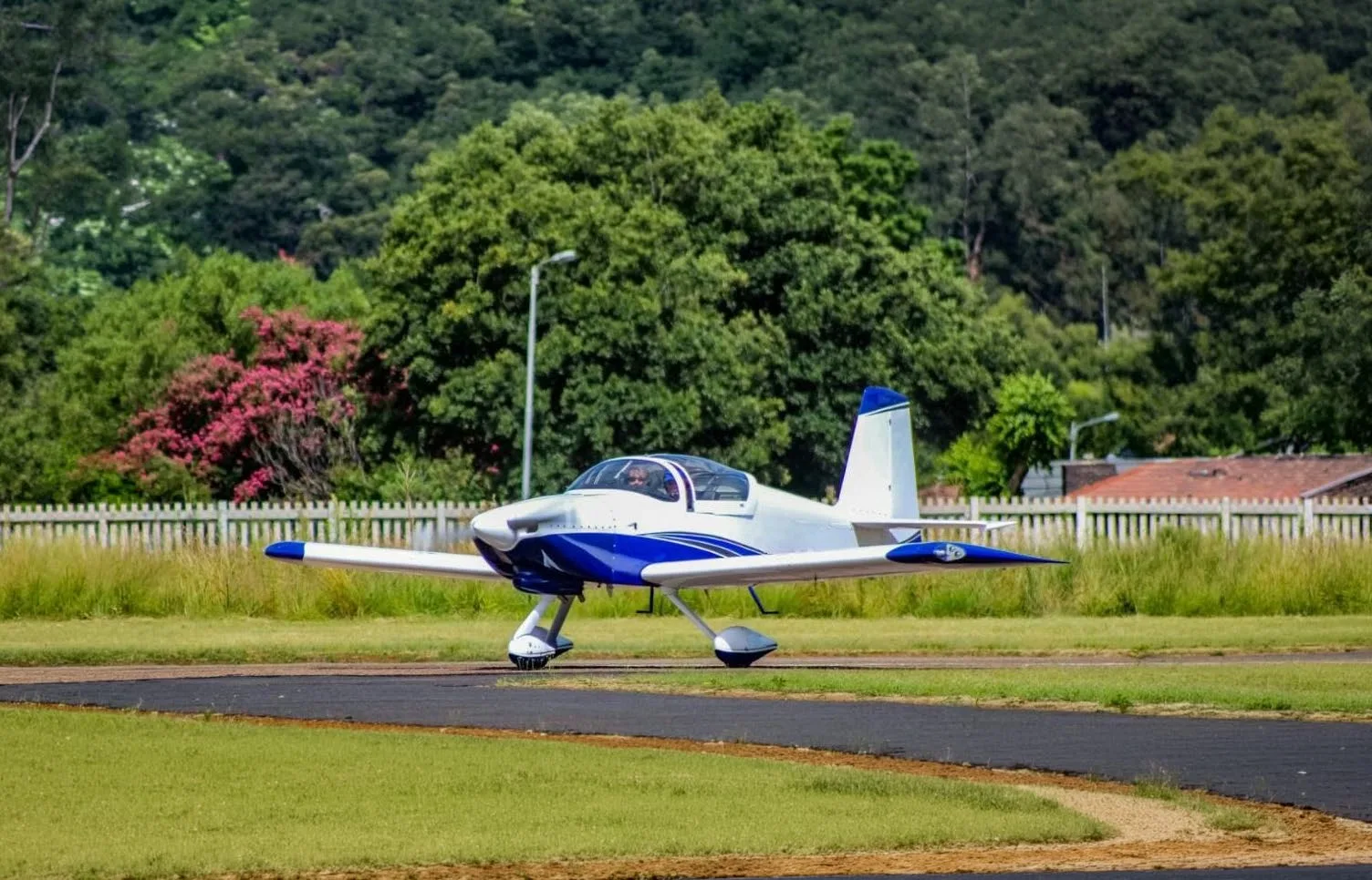 A white and blue single-engine aircraft with a bubble canopy on a runway, with green trees in the background.