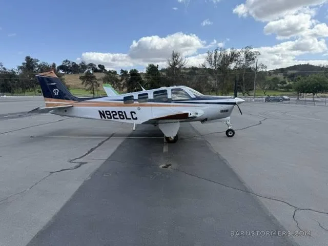 A white, blue, and copper Beechcraft Bonanza N926LC parked on a tarmac under a partly cloudy sky.