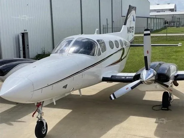 A white twin-engine Cessna 414 Chancellor aircraft with black and gold stripes, parked on an tarmac in front of a hangar, showing the registration N41019 on the tail.