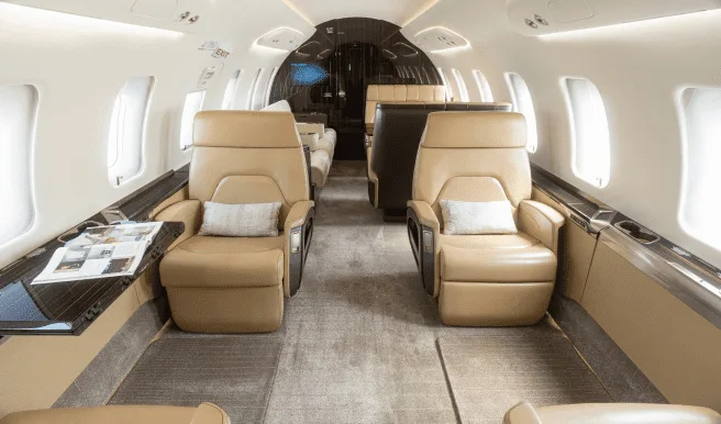 A view down the aisle of a private jet cabin featuring tan leather seats, a light-colored carpet, and overhead lighting.
