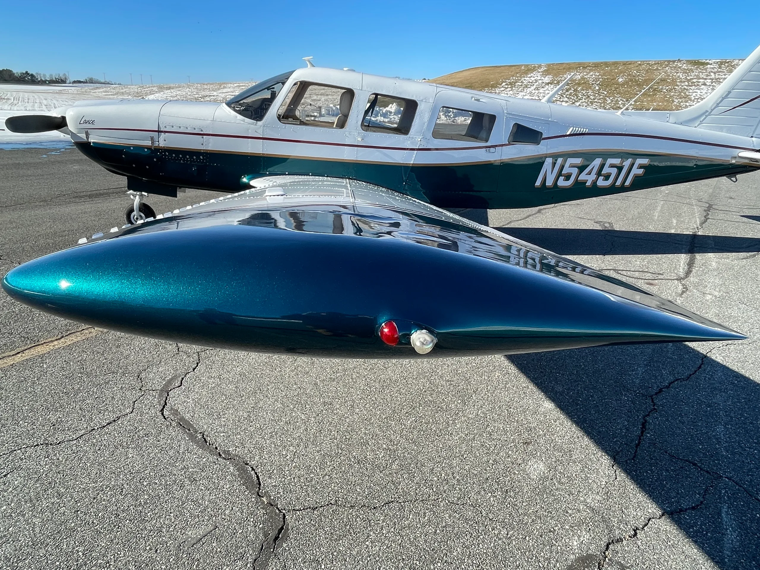 A white and dark green Piper Lance aircraft, registration N5451F, parked on an asphalt ramp with a snowy field in the background.