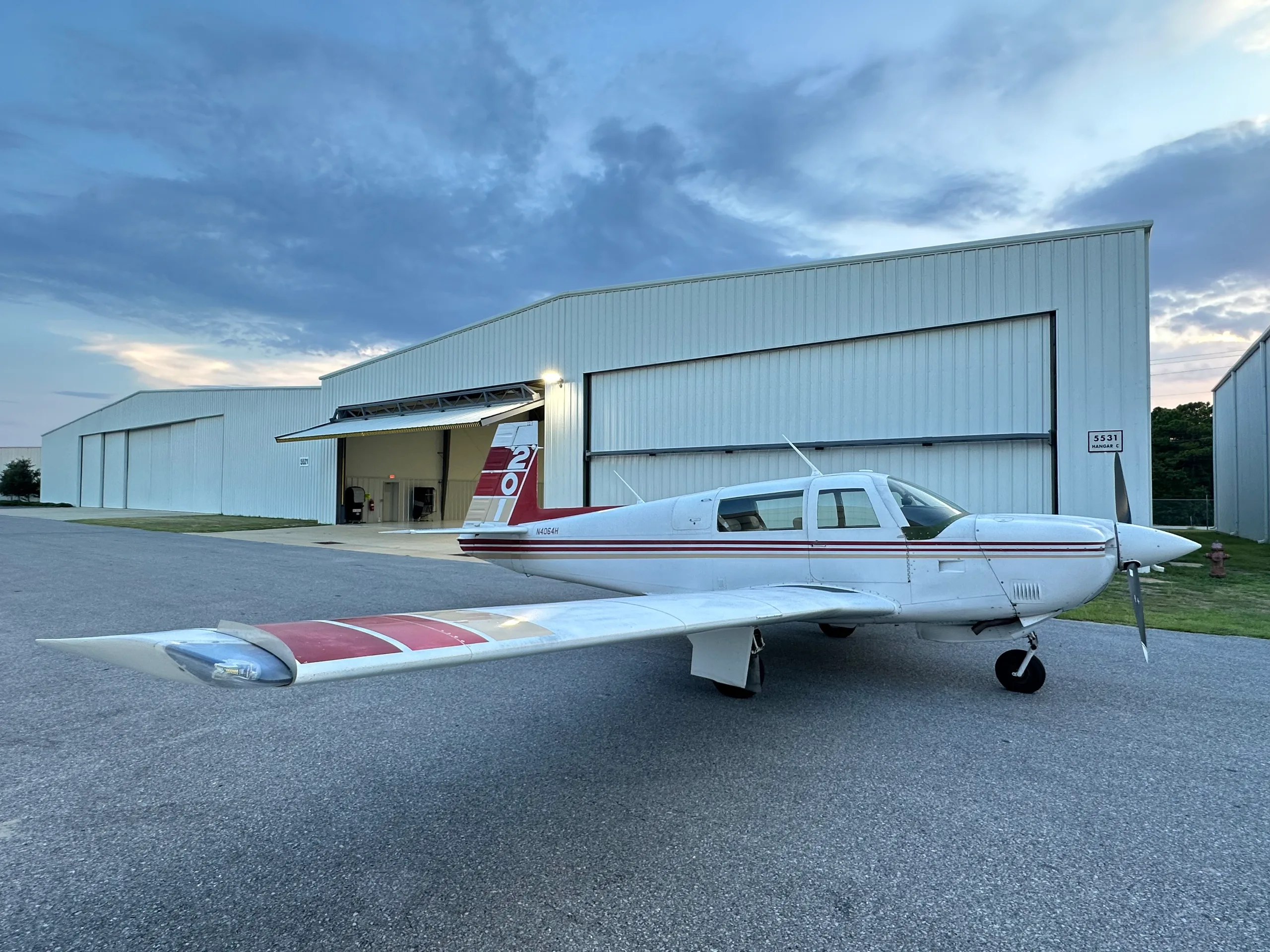 A white single-engine aircraft with red and gold stripes on the tail and wings, parked on an asphalt apron outside a hangar at dusk.