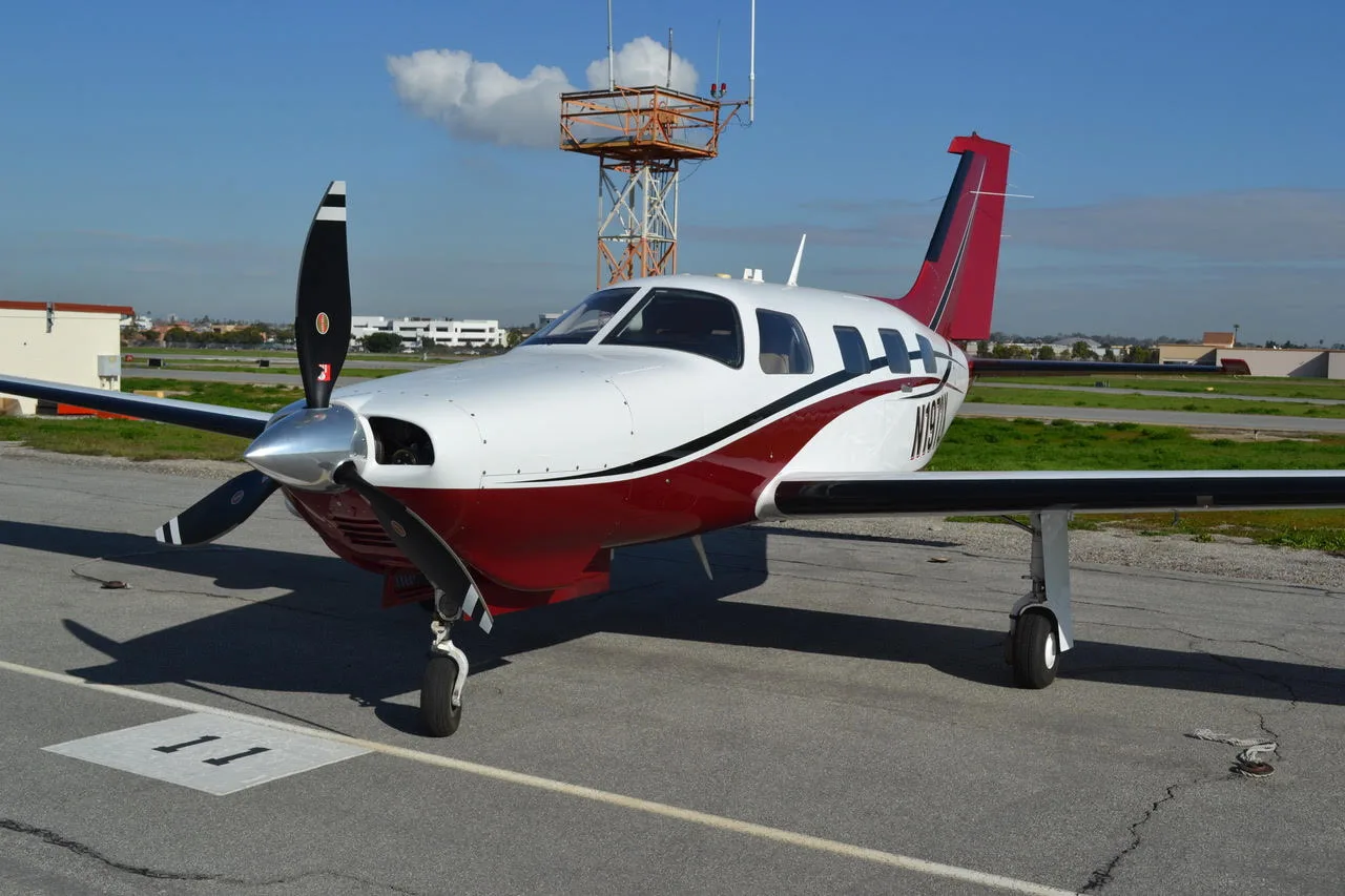 A white and red single-engine propeller aircraft, possibly a Piper Malibu, is parked on an airfield ramp with a tower in the background.
