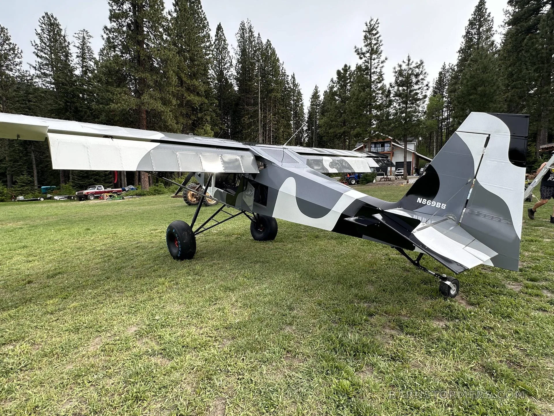 A black, white, and gray camouflage STOL aircraft with large tundra tires is parked on a grassy field, viewed from the rear-left.