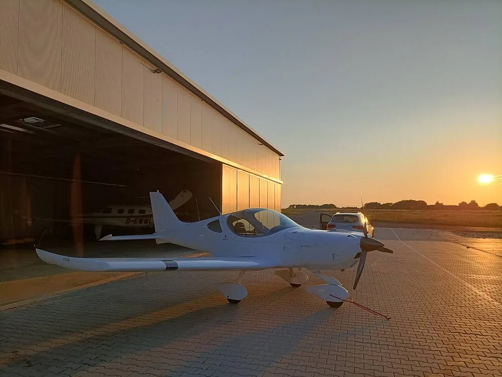 A white Bristell aircraft parked outside a hangar with the sun setting in the background.