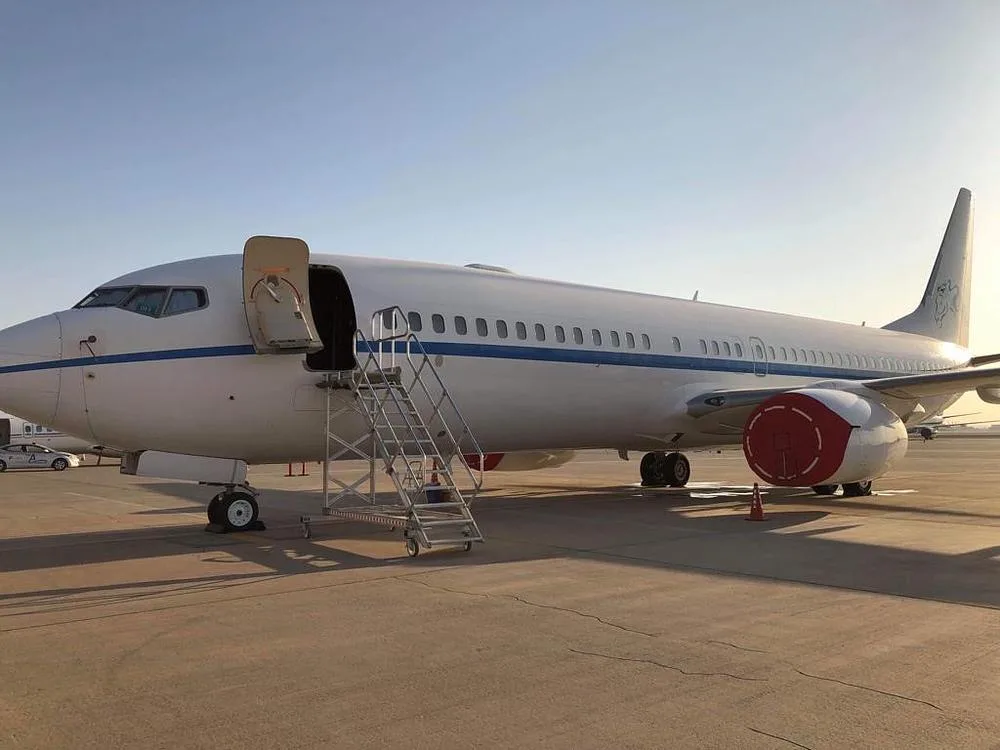 A white Boeing 737 with a blue stripe and red engine covers is parked on an airfield with boarding stairs.