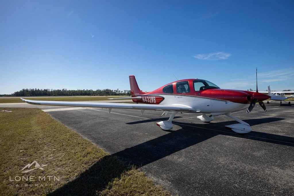 A red and white Cirrus SR20 aircraft (N432WB) parked on an airport tarmac under a clear blue sky.