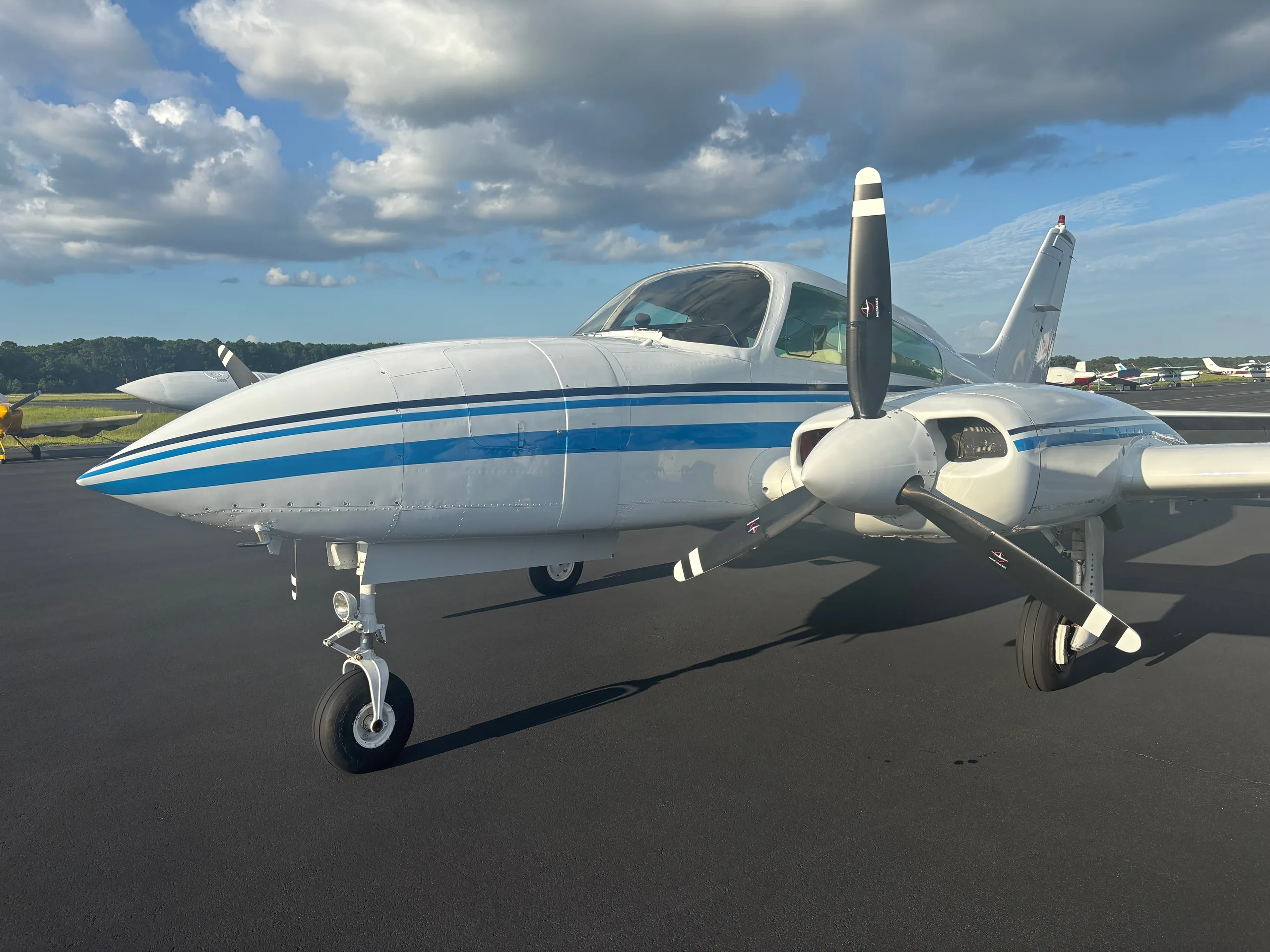 A white twin-engine aircraft with blue stripes parked on an asphalt runway, viewed from the front-left.