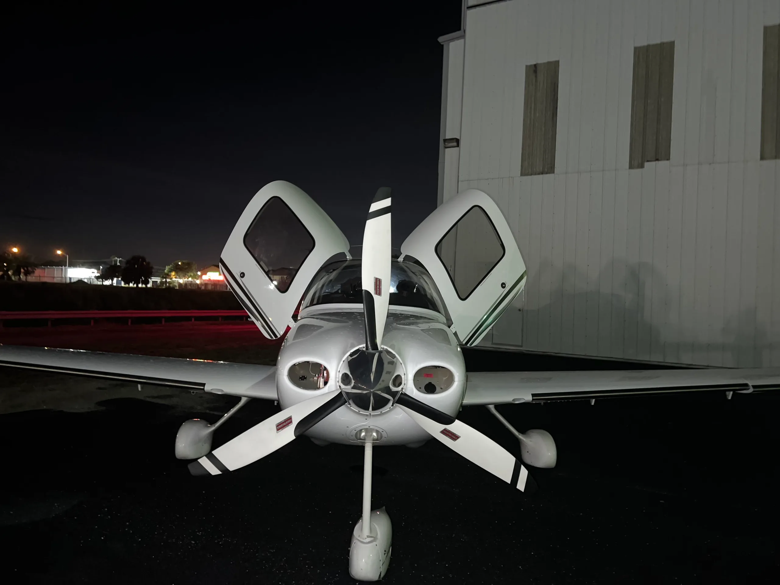 A white Cirrus SR22T aircraft with its gull-wing doors open, parked at night in front of a hangar.