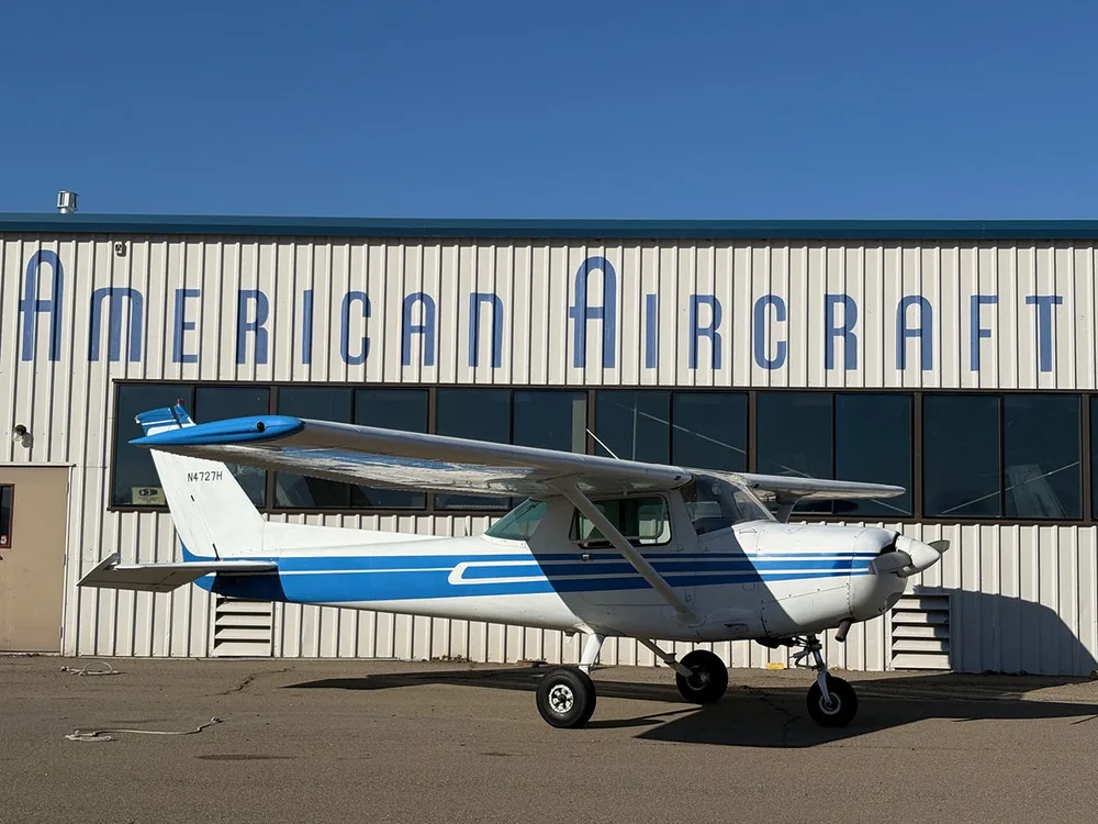 A white and blue Cessna 172 aircraft (N4727H) parked on an airfield in front of a hangar.