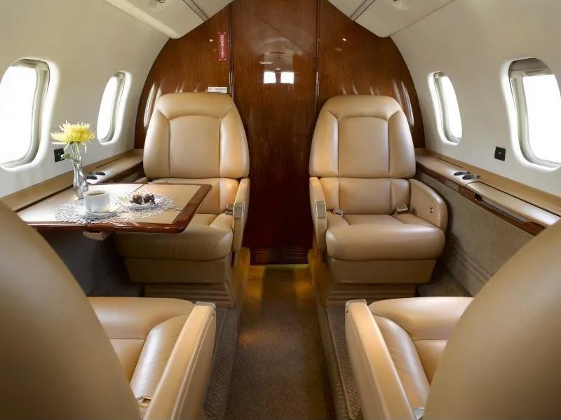 The luxurious tan leather interior of a private jet cabin, featuring two forward-facing seats and a table with refreshments.