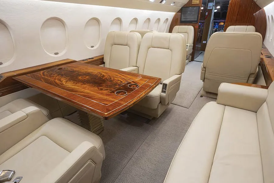 The interior of a private jet cabin, showing cream-colored leather seats and a polished wooden table extended for use.