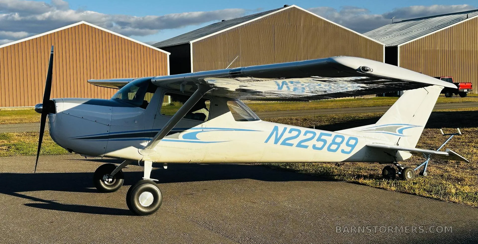 A white Cessna 150/152 type aircraft with blue and grey stripes, tail number N22589, parked on an asphalt ramp with hangars in the background.