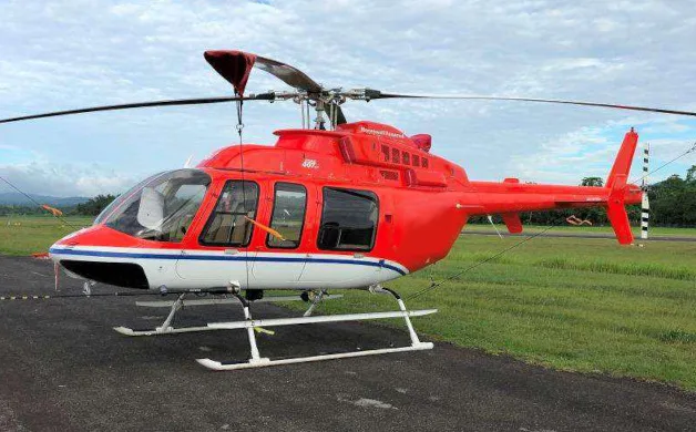 A red and white Bell 407HP helicopter with blue stripes is parked on an airfield under a cloudy sky.