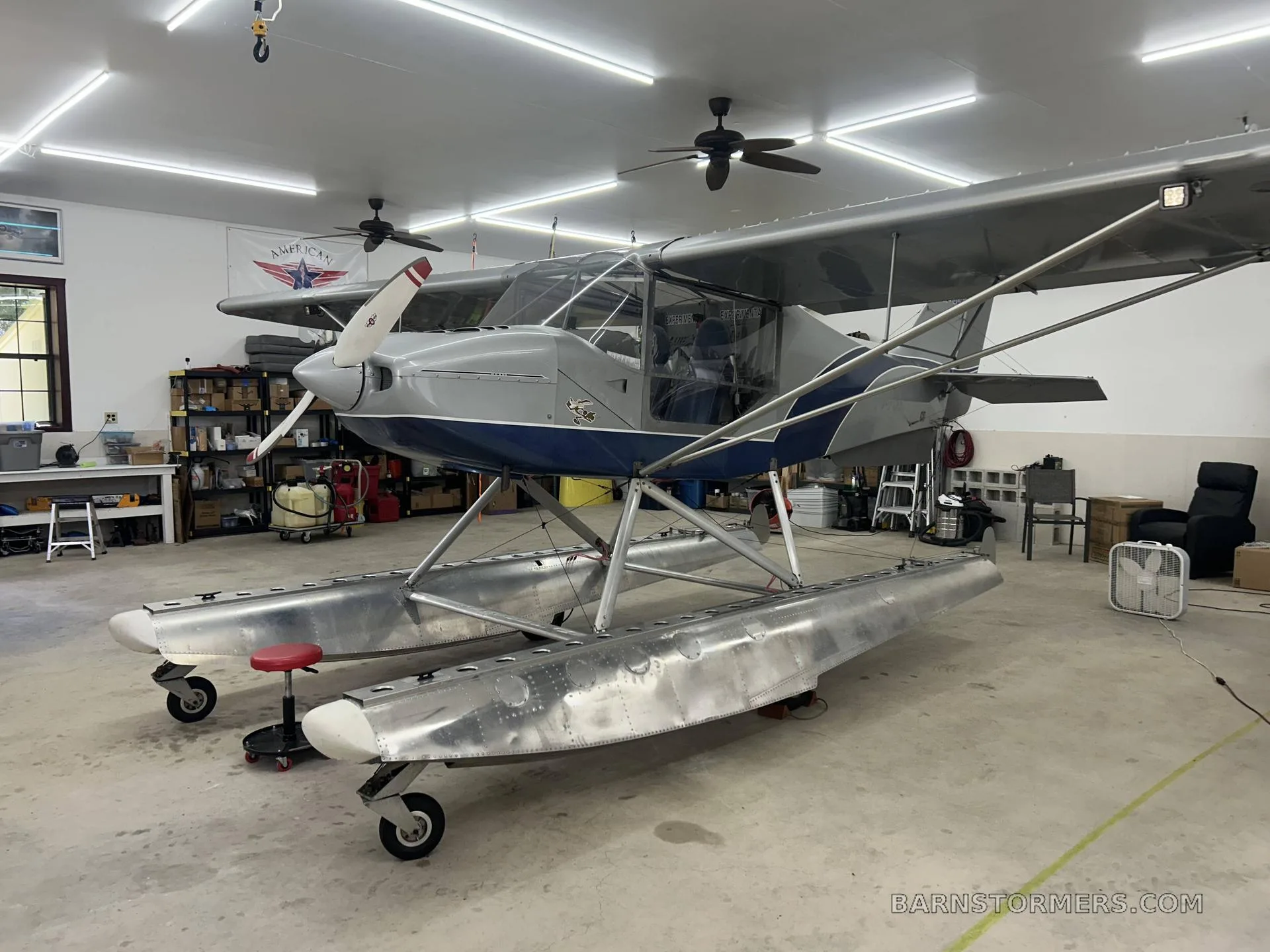 A silver and blue floatplane with a single propeller is parked inside a hangar.