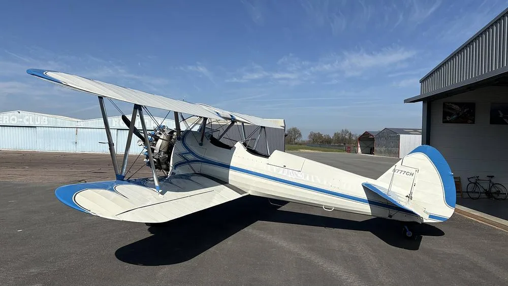 A white and blue biplane, N777CH, parked on an airfield next to a hangar on a sunny day.