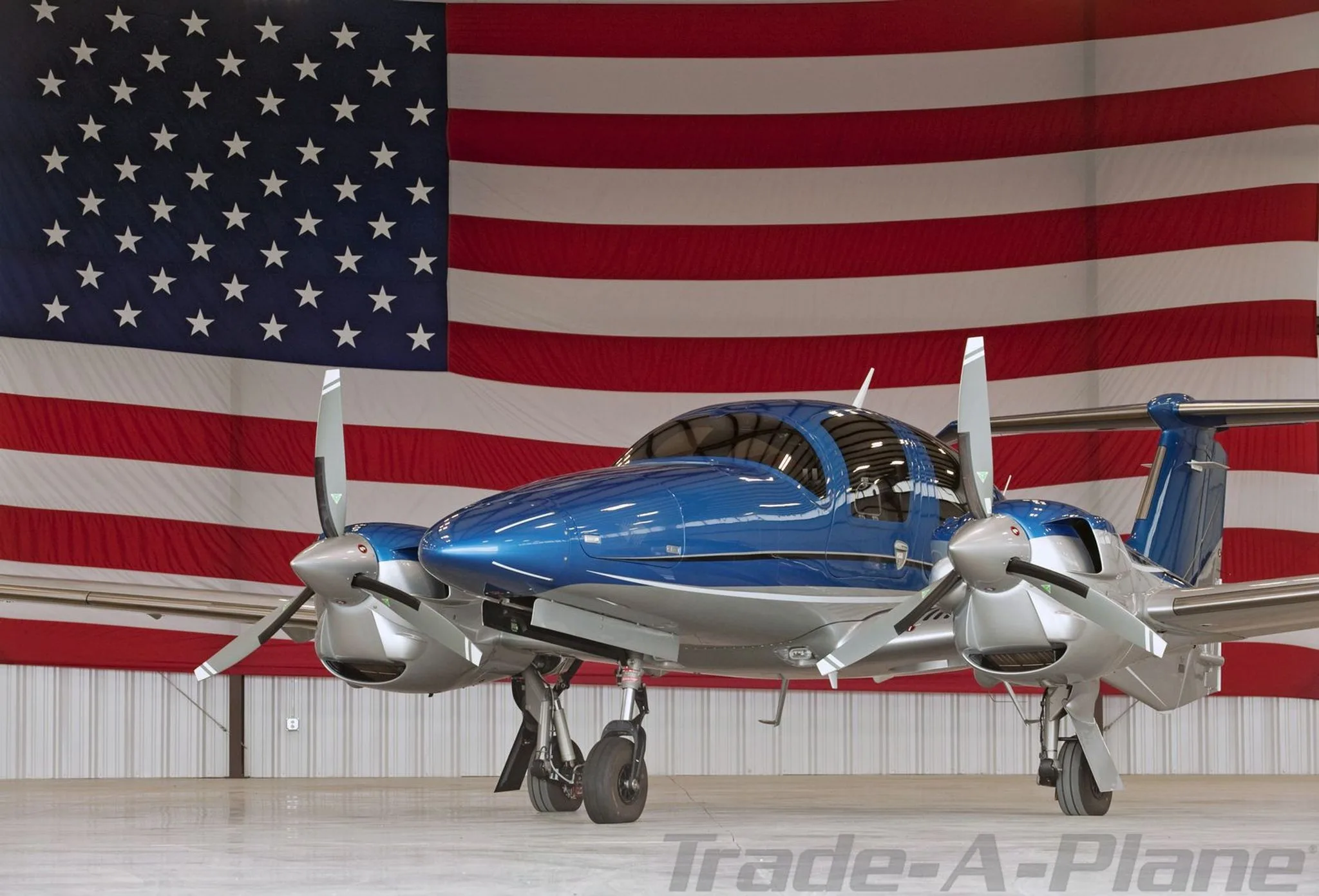 A blue and silver Diamond DA62 aircraft parked in a hangar with a large American flag in the background.