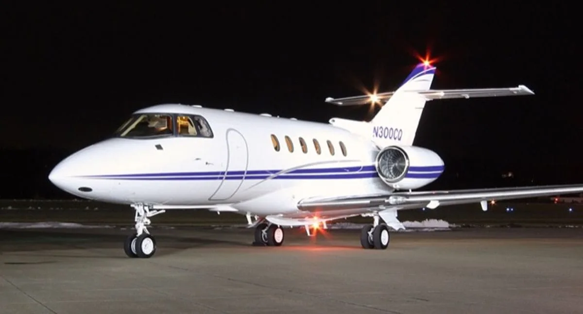 A white business jet with blue stripes and registration N300CO is parked on an airfield at night, illuminated by lights.