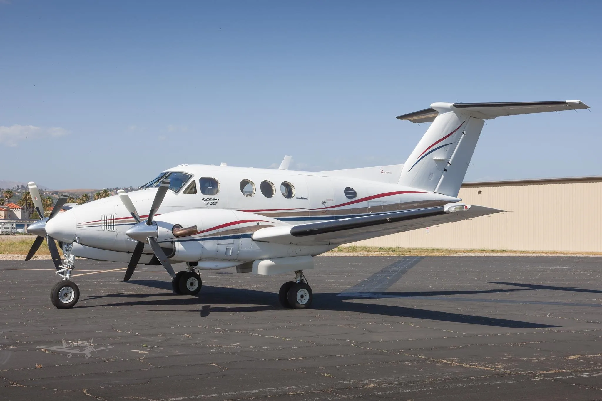A white King Air F90 with red and blue stripes is parked on an airport tarmac under a clear sky.