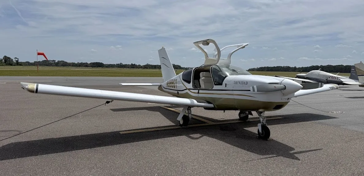 A white and gold Socata TB-20 Trinidad aircraft with its doors open is parked on an airfield next to another aircraft.