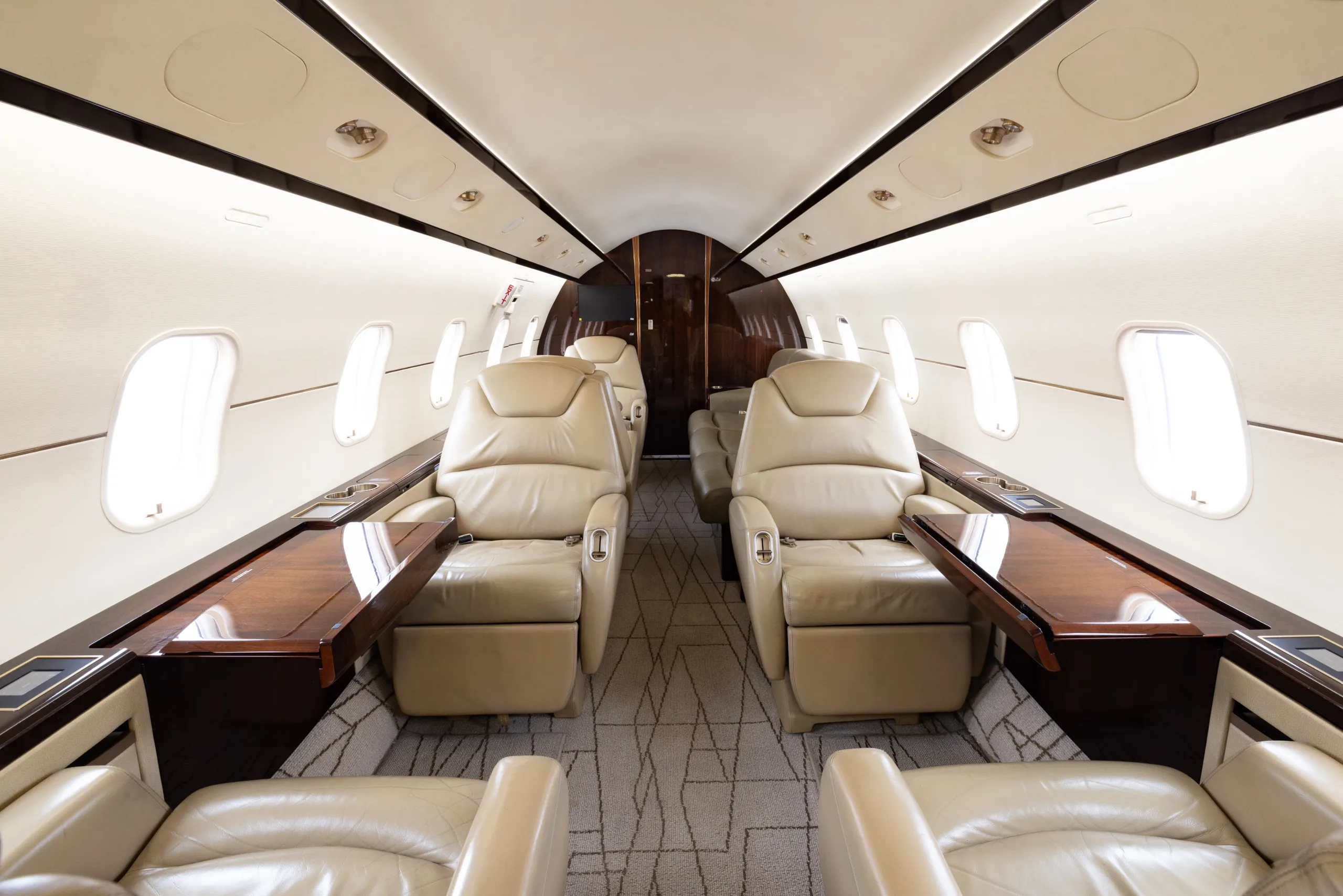 The luxurious interior of a business jet cabin with light beige leather seats and dark wood tables.