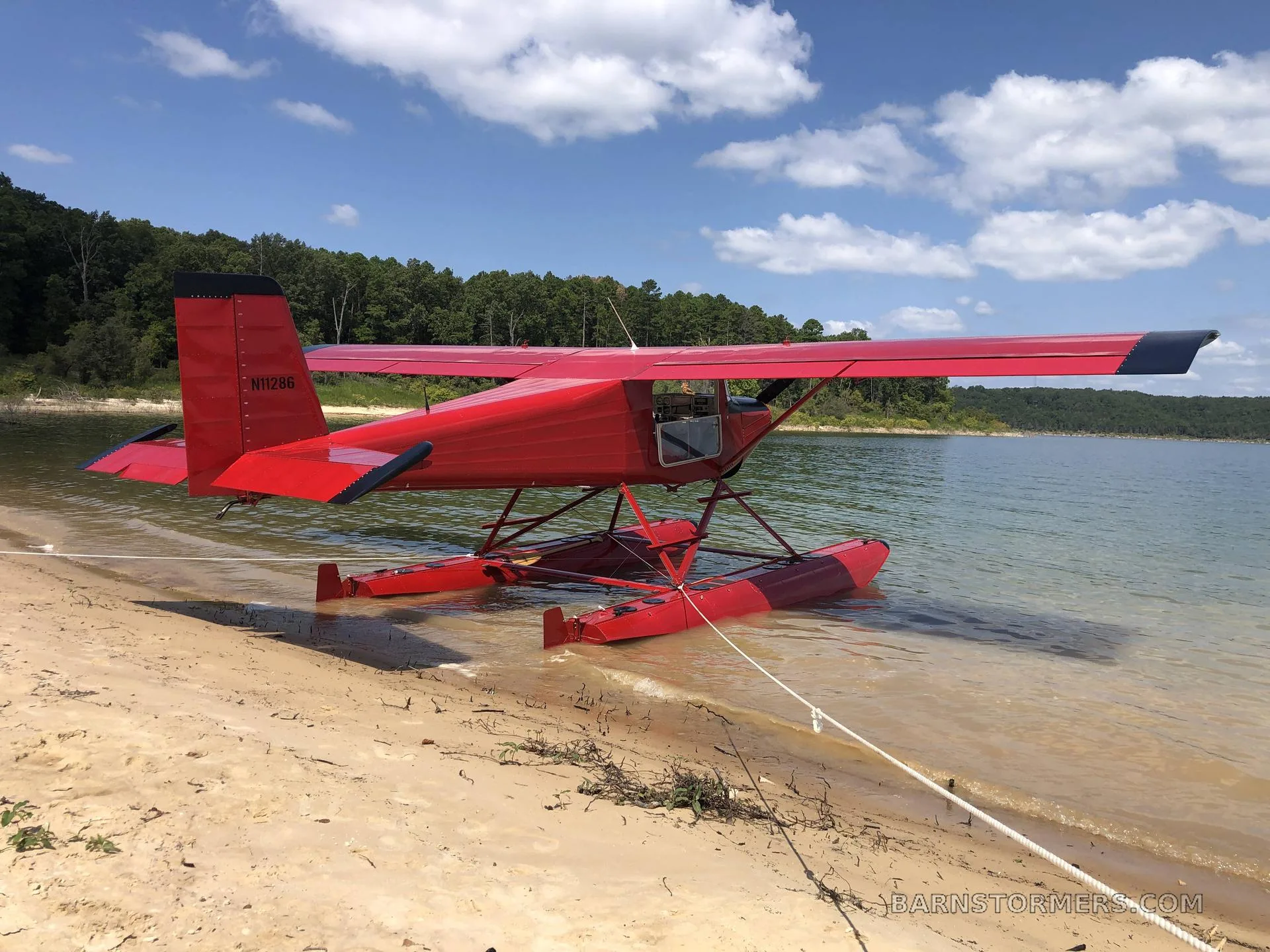 A red floatplane with registration N11286 is tied to a sandy beach, partially submerged in a lake under a blue sky.