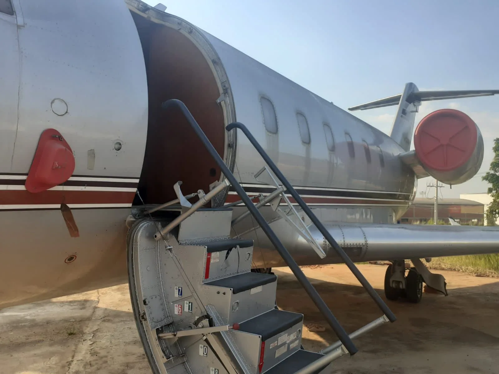 A side view of a silver business jet with open airstairs and a red engine cover, parked on a concrete surface.