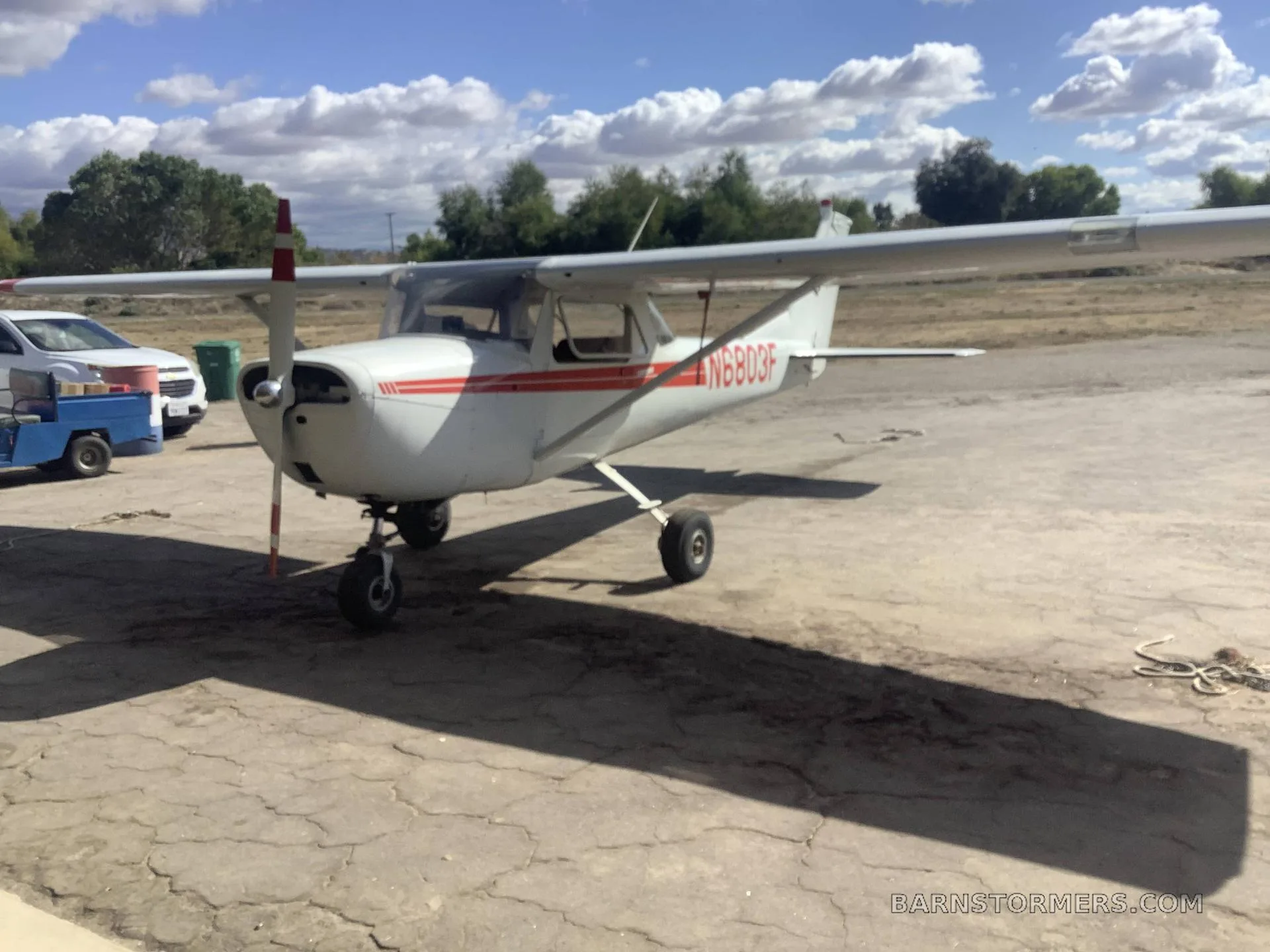 A white Cessna 150 with red stripes and registration N6803F is parked on a dirt-like surface under a partly cloudy sky.