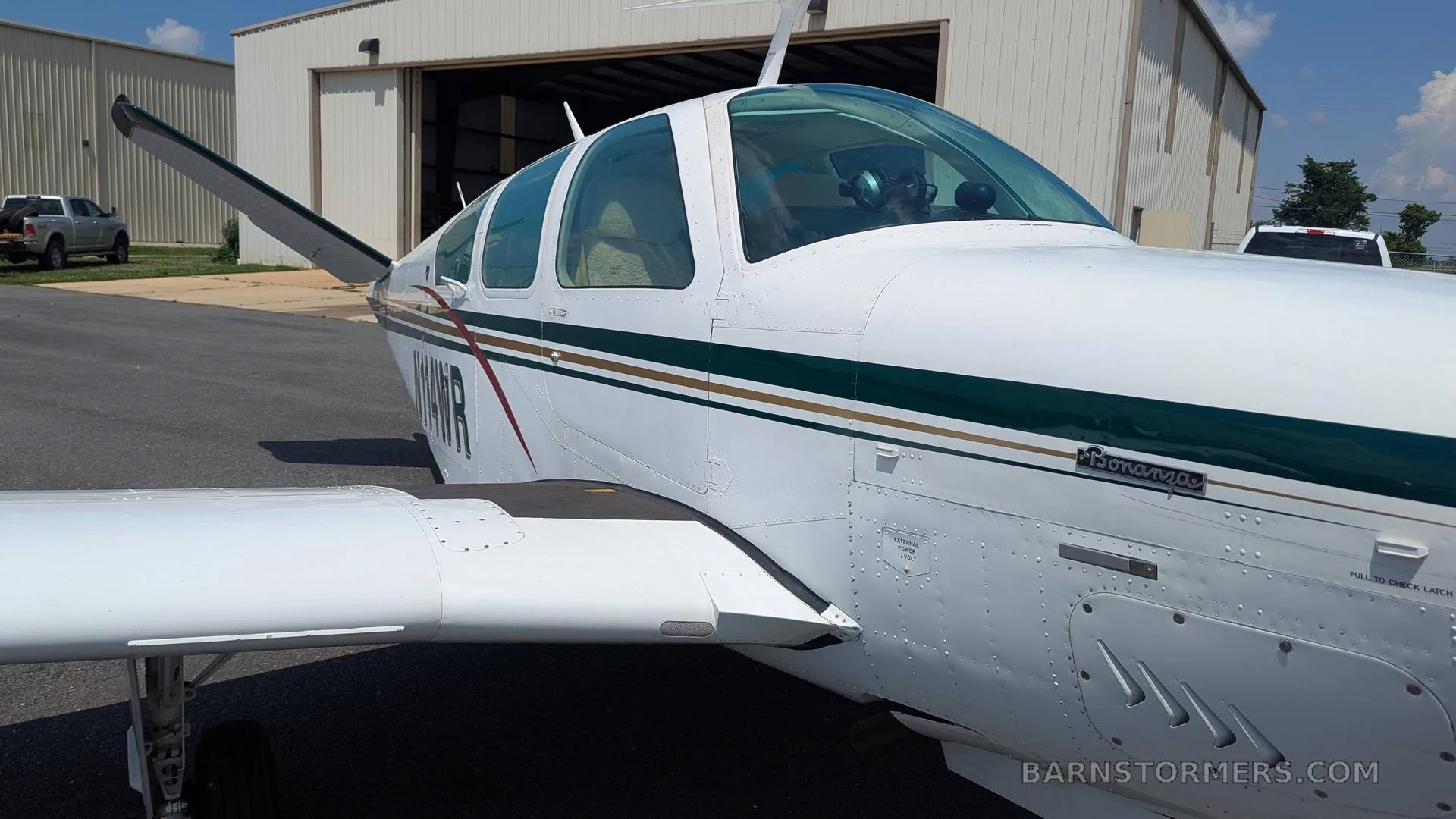 A white Beechcraft Bonanza with green and gold stripes, registration N114WR, parked on an asphalt ramp in front of a hangar.