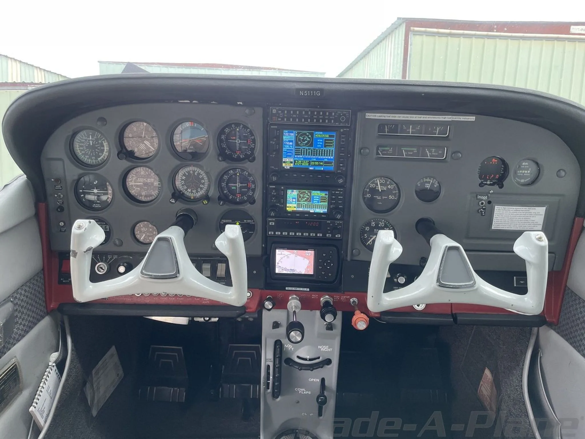 The cockpit of a Cessna 182 aircraft, showing the instrument panel with analog gauges and digital displays, and two white control yokes.