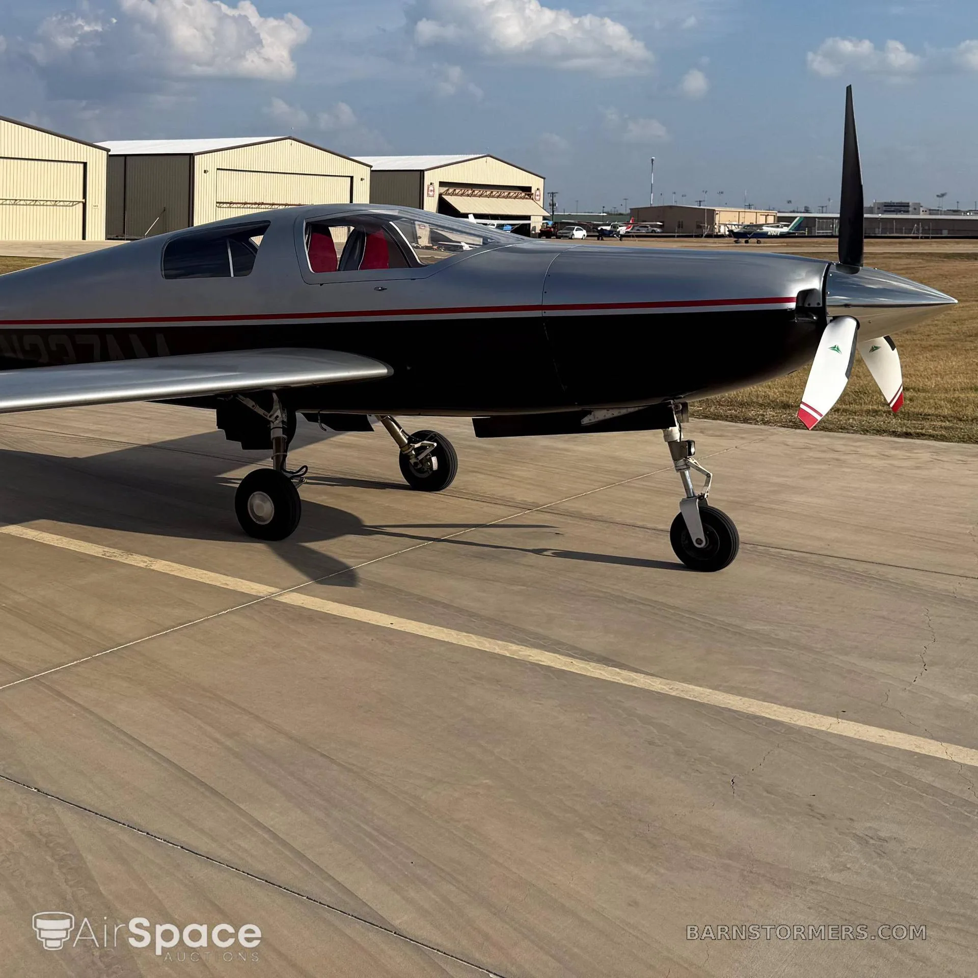 A silver and black single-engine aircraft with a red stripe, parked on an asphalt ramp with hangars in the background.