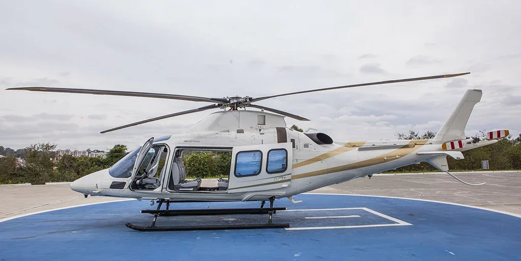 A white helicopter with gold stripes is parked on a blue helipad with its cabin doors open, viewed from the side.