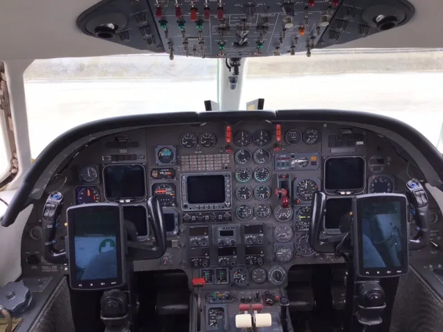 The cockpit of a business jet displays a complex instrument panel with numerous analog gauges, digital screens, and an overhead panel.