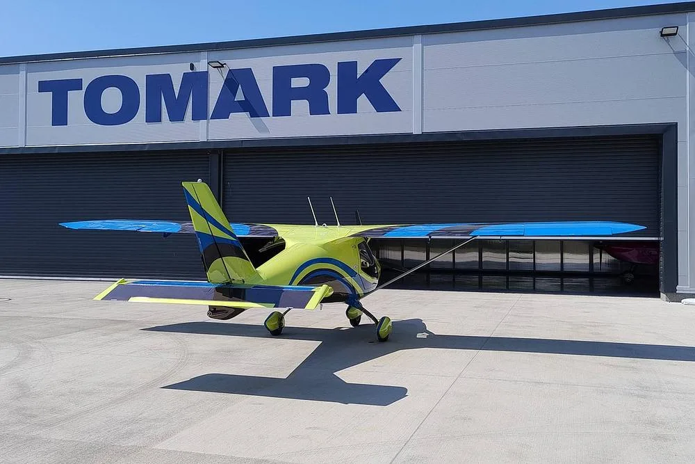 A yellow and blue Skyper GT9 aircraft parked on an airfield in front of a large hangar with 'TOMARK' written on it.
