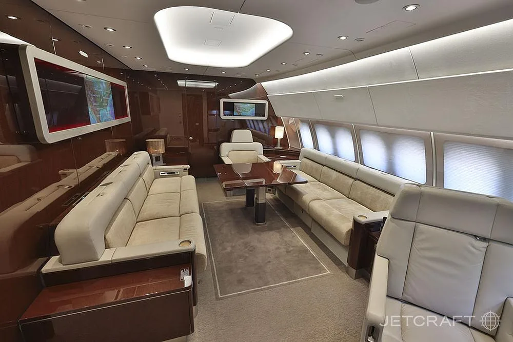 A spacious, luxurious aircraft interior featuring cream sofas, a dining table, and multiple large display screens.